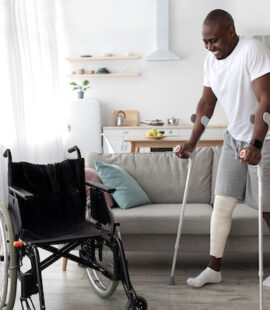 Surgery rehabilitation of bone and orthopaedic recovery, stay at home. Smiling adult african american guy with broken leg in cast walks with crutches to wheelchair in living room interior, panorama
