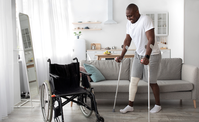 Surgery rehabilitation of bone and orthopaedic recovery, stay at home. Smiling adult african american guy with broken leg in cast walks with crutches to wheelchair in living room interior, panorama