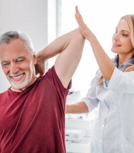 A female orthopedist or physical therapist assisting an arm stretch with an older male patient, a good candidate for red light therapy wraps for joint pain.