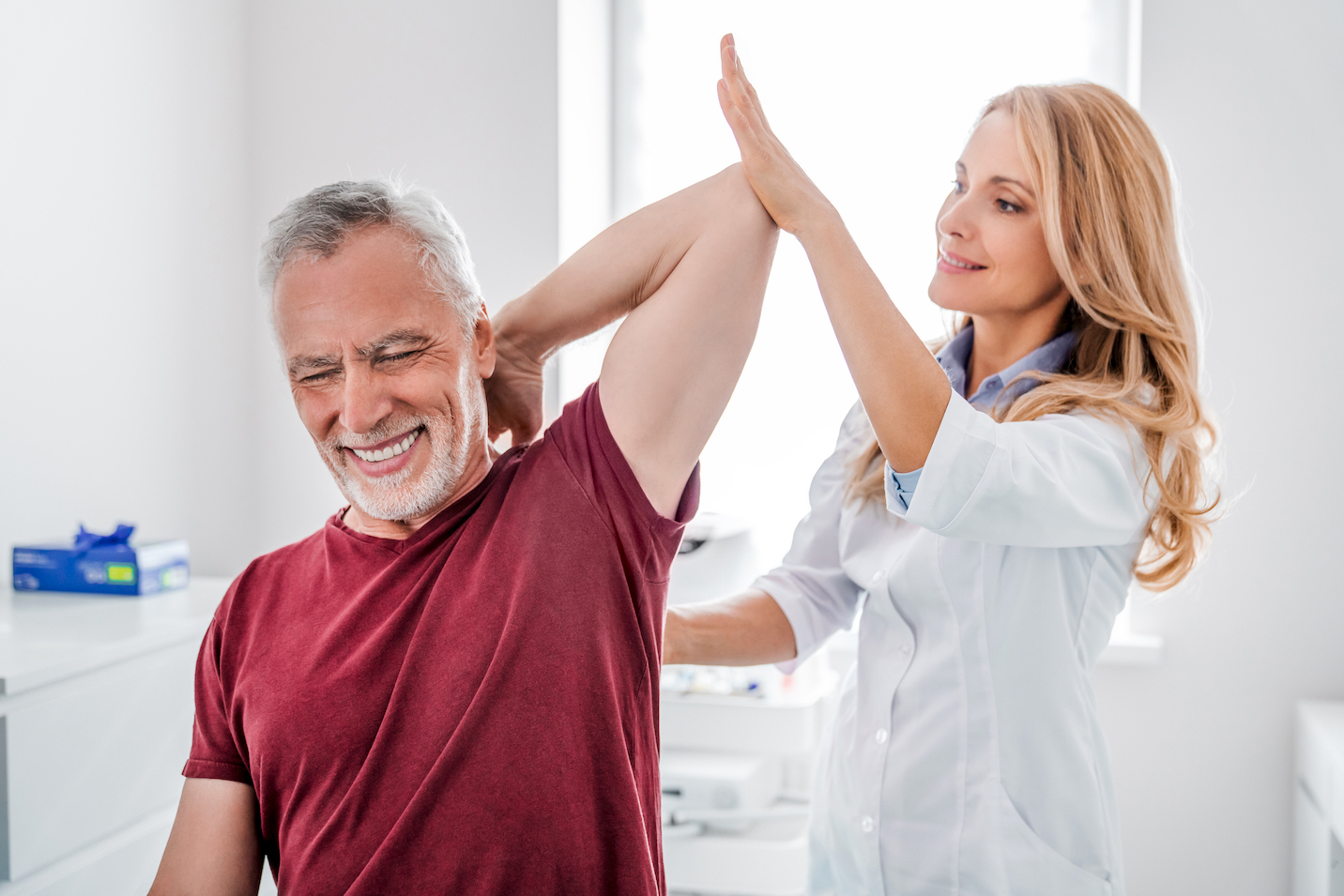 A female orthopedist or physical therapist assisting an arm stretch with an older male patient, a good candidate for red light therapy wraps for joint pain.