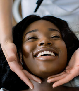 Close-up of hands of cosmetologist woman making special face massage to young pretty African smiling woman lying on couch in beauty salon. Concept of antiaging facial skin treatments and massage